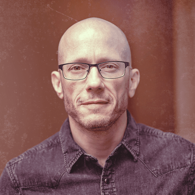 Jon Arnold, smiling slightly, in glasses and a dark denim shirt